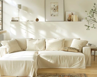 May include: A large, off-white sectional sofa with a linen-like fabric. The sofa has a chaise lounge on the right side and a large ottoman in front. The sofa is in a living room with a white wall and a rug on the floor.