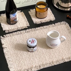 May include: Two square off-white fabric coasters with frayed edges and one rectangular off-white fabric placemat with frayed edges. The textile items feature a textured weave and are displayed on a dark surface alongside a bottle, candle, jar, and pitcher.
