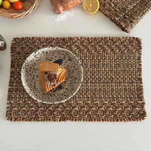 May include: A rectangular woven placemat in shades of brown and tan with a textured, grid-like pattern and frayed edges. A speckled ceramic bowl sits on the placemat, containing a slice of layered cake topped with a rectangular cookie.