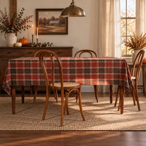 May include: A rectangular dining table covered with a red, black, and white plaid tablecloth. The table is surrounded by four wooden chairs with curved backrests and woven seats. The setup is placed on a textured, light brown woven area rug.