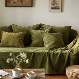 以下が含まれることがあります： 緑色のベルベットのソファと、お揃いの正方形のクッションとスロー。木製のコーヒーテーブルには、白い花の花瓶と2つのティーカップが置かれています。温かみのあるリビングルームの雰囲気。