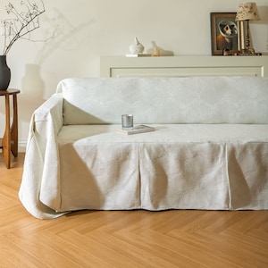 Op de afbeelding: Een witte bank met een witte, geborduurde slipcover. De bank heeft een rok en staat op een houten vloer.