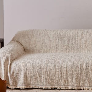 May include: A cream-colored sofa cover with a textured, shaggy fabric surface. The cover features a straight hemline along the bottom edge finished with a short, matching fringe detail that runs across the entire width of the furniture piece.