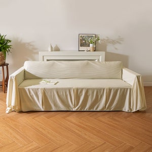 Puede incluir: Un sofá blanco con una funda a rayas beige y blanca. El sofá está sobre un suelo de madera con un patrón de espiga.