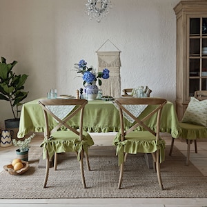 以下が含まれることがあります： 緑色のリネン製のテーブルクロスが、フリルのある縁取りで、4脚の椅子のある木製ダイニングテーブルを覆っています。椅子には、フリルのある緑色のリネン製のクッションと、クロスバックのデザインが施されています。テーブルの上には、青い花が飾られた花瓶と、グラスなどの装飾品が置かれています。