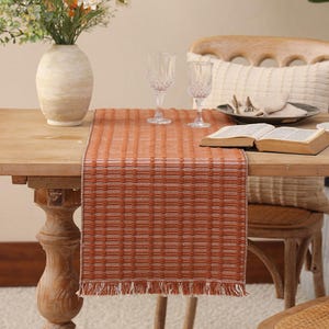 May include: A rectangular table runner featuring a woven pattern of burnt orange and white threads. The fabric has a textured, grid-like appearance with a decorative fringe along the bottom edge, creating a distinct contrast against the wooden table.