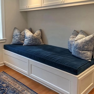 May include: A white built-in bench with a long, dark blue velvet cushion and four square throw pillows featuring a blue and white toile pattern. The bench base includes two rectangular cabinet panels, and white cabinets are mounted above.