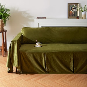 Puede incluir: Una funda de sofá de terciopelo verde oscuro con una falda plisada. La funda está drapeada sobre un sofá con una superficie blanca detrás.