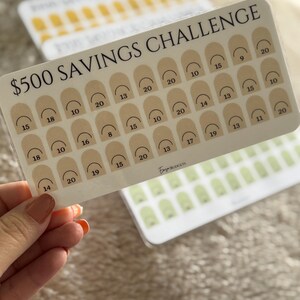 Op de afbeelding: Een doorzichtige plastic spaarchallengekaart met de tekst "$500 SAVINGS CHALLENGE" in het zwart. De kaart heeft een raster van beige bogen met cijfers. Extra kaarten met vergelijkbare ontwerpen zijn zichtbaar op de achtergrond.