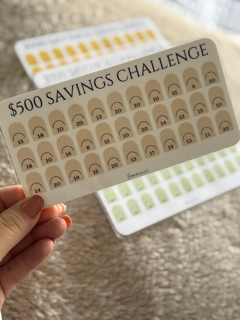 Op de afbeelding: Een doorzichtige plastic spaarchallengekaart met de tekst "$500 SAVINGS CHALLENGE" in het zwart. De kaart heeft een raster van beige bogen met cijfers. Extra kaarten met vergelijkbare ontwerpen zijn zichtbaar op de achtergrond.
