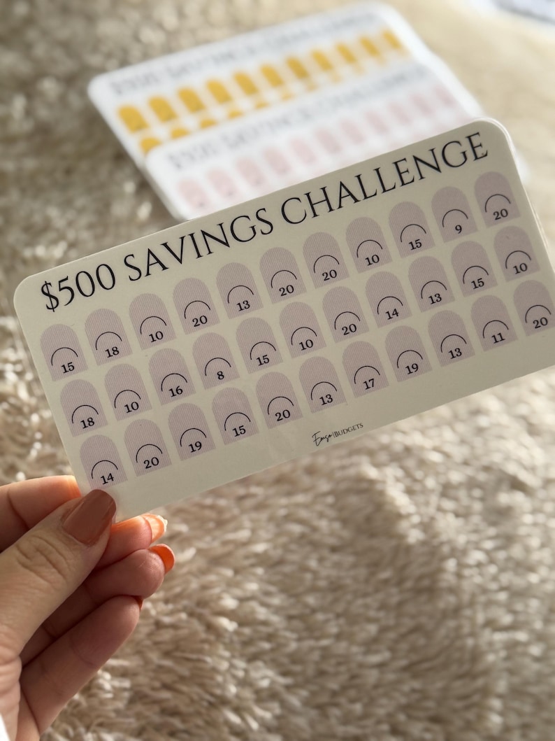 Op de afbeelding: Een spaaruitdagingskaart met de tekst "$500 SAVINGS CHALLENGE" in het zwart. De kaart heeft een lichtbeige achtergrond met rijen boogvormen, elk met een nummer erin. De kaart wordt door een persoon vastgehouden.