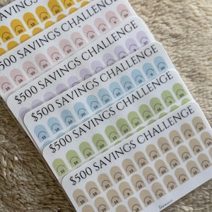 Op de afbeelding: Een stapel kleurrijke sticker vellen met de tekst "$500 SAVINGS CHALLENGE". Elke vel bevat rijen genummerde bogen in tinten geel, roze, paars, blauw, groen en beige. De nummers vari&euml;ren van 9 tot 20.
