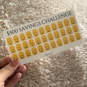Op de afbeelding: Een witte spaaruitdagingskaart met de tekst "$500 SAVINGS CHALLENGE" in het zwart. De kaart heeft gele boogvormen met cijfers erin. De kaart wordt door een persoon vastgehouden.