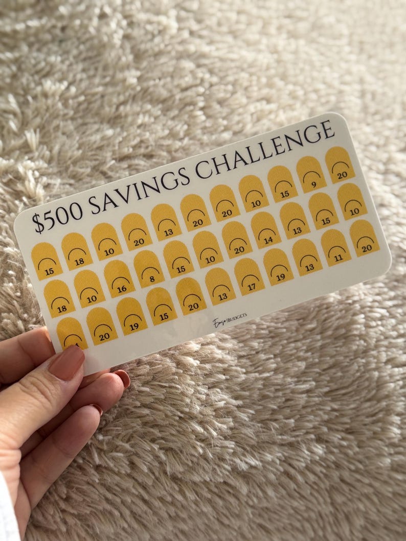 Op de afbeelding: Een witte spaaruitdagingskaart met de tekst "$500 SAVINGS CHALLENGE" in het zwart. De kaart heeft gele boogvormen met cijfers erin. De kaart wordt door een persoon vastgehouden.