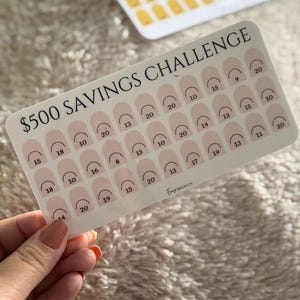 Op de afbeelding: Een witte spaarchallengekaart met de tekst "$500 SAVINGS CHALLENGE" en vakken om te sparen bij te houden. De kaart heeft een roze en wit ontwerp met nummers die spaarbedragen aangeven. Een tweede kaart is zichtbaar op de achtergrond.