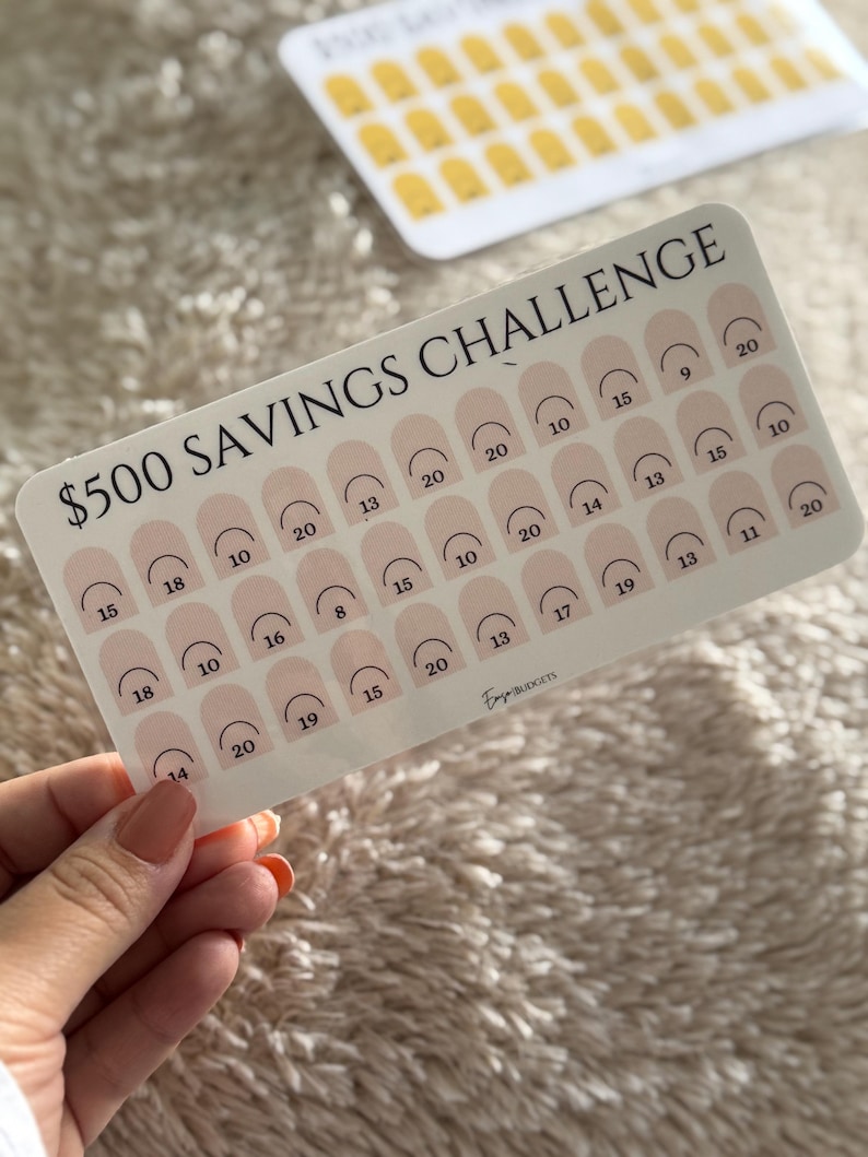 Op de afbeelding: Een witte spaarchallengekaart met de tekst "$500 SAVINGS CHALLENGE" en vakken om te sparen bij te houden. De kaart heeft een roze en wit ontwerp met nummers die spaarbedragen aangeven. Een tweede kaart is zichtbaar op de achtergrond.