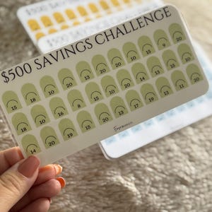 Op de afbeelding: Een witte spaarchallengekaart met de tekst "$500 SAVINGS CHALLENGE" in het zwart. De kaart heeft een raster van groene vormen met daarin cijfers. De kaart wordt door een persoon vastgehouden.