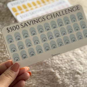 Op de afbeelding: Een spaaruitdagingskaart met de tekst "$500 SAVINGS CHALLENGE" en een raster van genummerde vakken. De kaart wordt in een hand gehouden. Extra kaarten zijn zichtbaar op de achtergrond. De kaart heeft een lichtbeige achtergrond met lichtblauwe bogen.
