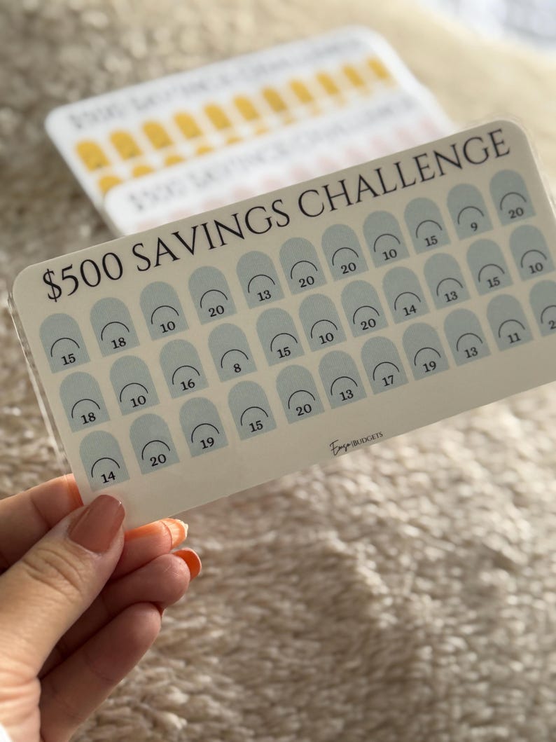 Op de afbeelding: Een spaaruitdagingskaart met de tekst "$500 SAVINGS CHALLENGE" en een raster van genummerde vakken. De kaart wordt in een hand gehouden. Extra kaarten zijn zichtbaar op de achtergrond. De kaart heeft een lichtbeige achtergrond met lichtblauwe bogen.