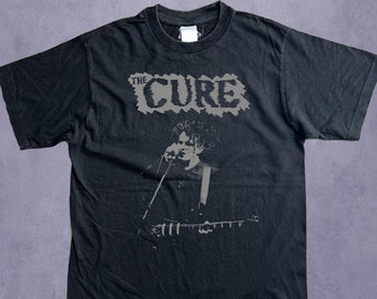 May include: Black t-shirt with a grey graphic of The Cure. The band's name is printed above a grayscale image of a person playing guitar and singing. The shirt has a crew neck and is made of a dark fabric.