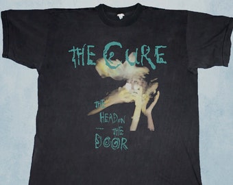 May include: Black t-shirt with the band name "The Cure" in teal, above a blurred image. Below the image, the text reads "The Head on the Door" in teal. A vintage band tee.