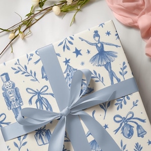 Peut inclure: Un coffret cadeau emballé avec un ruban de satin bleu clair noué en nœud. Le papier d'emballage présente un motif de toile de Jouy bleu et blanc avec des ballerines, des casse-noisettes et des sapins de Noël. Le cadeau est sur une surface en marbre blanc.