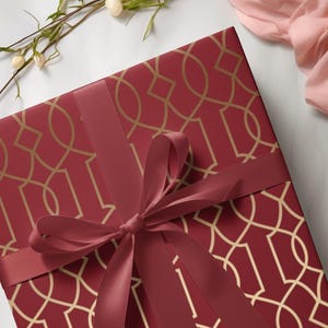 May include: A square gift box wrapped in burgundy paper with a gold geometric pattern. A burgundy ribbon is tied in a bow on top. The box is on a white surface with flowers and pink fabric.