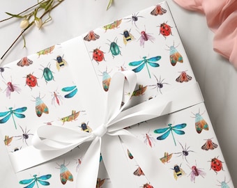 Puede incluir: Una caja de regalo cuadrada envuelta en papel blanco con un patrón colorido de varios insectos, incluyendo mariquitas, libélulas, abejas y arañas. La caja está atada con una cinta de satén blanca dispuesta en un lazo.