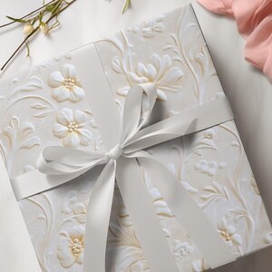 Peut inclure: Un coffret cadeau carr&eacute; avec un motif floral en relief de couleur cr&egrave;me, nou&eacute; avec un ruban de satin blanc. La bo&icirc;te pr&eacute;sente un motif r&eacute;p&eacute;t&eacute; de fleurs et de feuilles. Le ruban est nou&eacute; en n&oelig;ud.