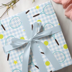 Puede incluir: Una caja de regalo de vichy azul y blanco con una cinta azul. La caja está decorada con un patrón de raquetas de tenis y pelotas de tenis.