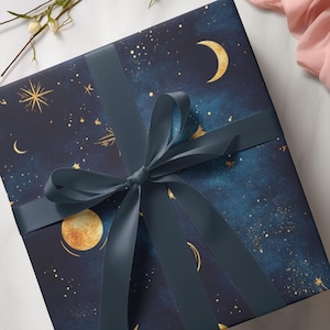 Könnte beinhalten: Ein verpacktes Geschenk mit marineblauem Papier mit einem Himmelsdesign aus goldenen Sternen, Monden und Planeten. Ein passendes marineblaues Band ist zu einer Schleife gebunden. Das Geschenk liegt auf einer weißen Marmoroberfläche.