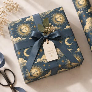 May include: A square gift box wrapped in dark blue paper featuring a celestial pattern of suns, moons, stars, and clouds. A dark blue satin ribbon is tied in a bow, accented with dried greenery and a tag reading A LITTLE MAGIC INSIDE.