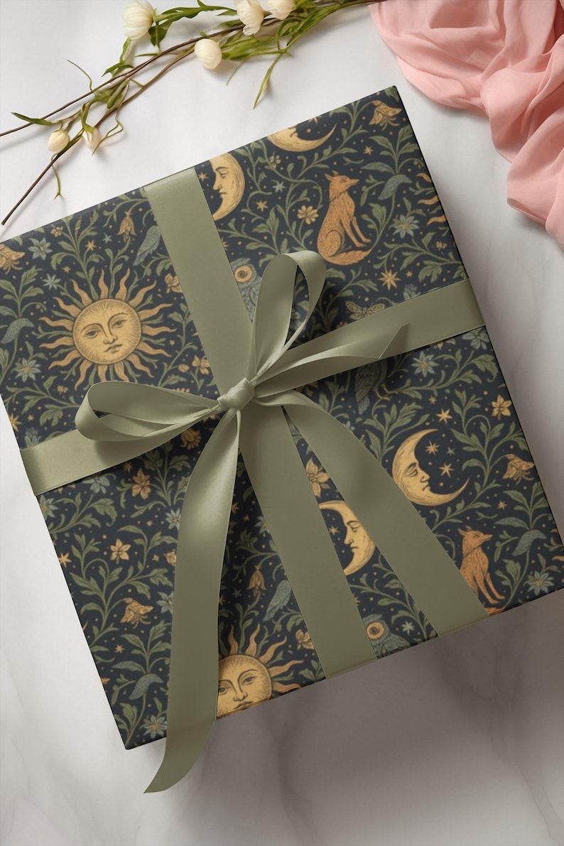 Puede incluir: Una caja de regalo envuelta en papel con un fondo azul oscuro y un patr&oacute;n repetido de soles, lunas, zorros y elementos florales. La caja est&aacute; atada con una cinta y un lazo verde salvia. El tema es celestial e inspirado en la naturaleza.