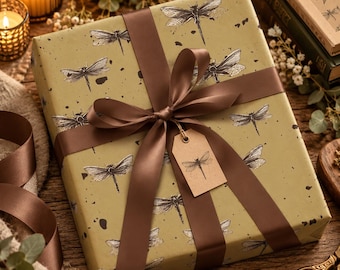 May include: A square gift box wrapped in olive green paper featuring a black dragonfly pattern and dark speckles. The box is tied with a wide brown satin ribbon and includes a small, rectangular kraft paper gift tag with a dragonfly illustration.