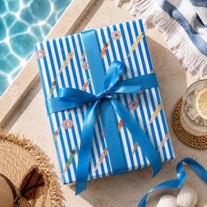 May include: A wrapped gift box with blue and white striped paper, adorned with a blue ribbon and bow. The wrapping paper features illustrations of swimmers and donuts. The scene includes a straw hat, sunglasses, and a drink with a lemon slice.