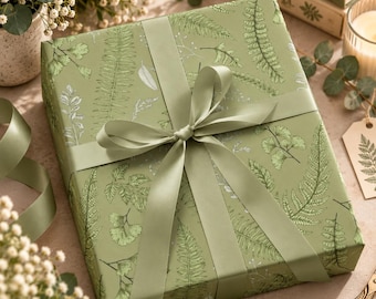Può includere: Una scatola regalo quadrata avvolta in carta verde salvia con una stampa botanica dettagliata di varie foglie di felce e fogliame in verde scuro e bianco. La scatola è legata con un ampio e liscio nastro di raso verde salvia.
