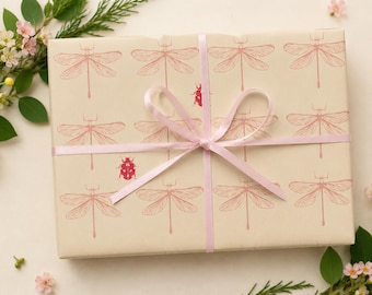 May include: A rectangular gift box wrapped in light beige paper featuring a repeating pattern of pink dragonfly illustrations and small red ladybug icons. The box is tied with a thin, light pink satin ribbon tied in a bow at the center.