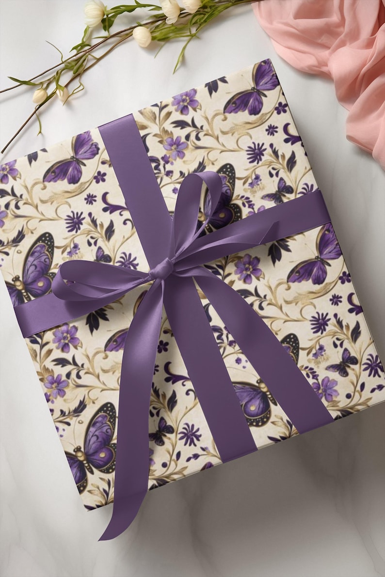 May include: Square gift box wrapped in cream-colored paper with a butterfly and floral pattern in purple and gold. A wide purple satin ribbon is tied in a bow around the box. The box is on a white marble surface.