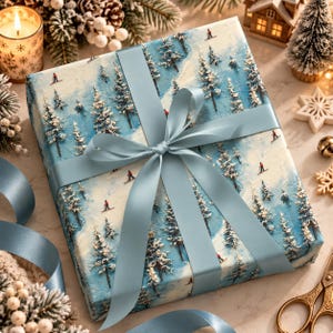May include: A square gift box wrapped in paper featuring a winter landscape with snow-covered pine trees and small figures of people skiing in red jackets. The box is tied with a wide, smooth, light blue satin ribbon in a centered bow.