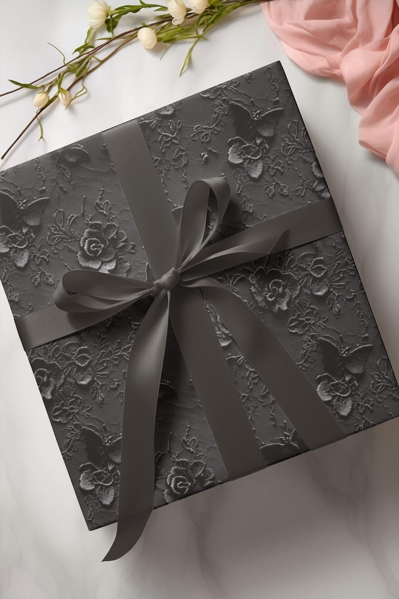 A square, charcoal-coloured gift box with a floral lace pattern and a matching satin ribbon tied in a bow. The box is decorated with embossed flowers and butterflies. The background includes white and pink fabric and white flowers.