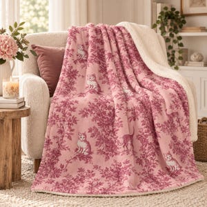 May include: A soft throw blanket featuring a pink and dark red toile pattern of cats and foliage. The blanket has a plush, cream-colored sherpa fleece lining on the reverse side, providing a textured contrast to the smooth, printed top surface.