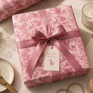 Peut inclure: Un cadeau emballé dans du papier rose avec un motif de chat et de fleurs, noué avec un ruban et un nœud roses. Une étiquette indique "Especially For You". Un rouleau de papier d'emballage assorti est visible. Des ciseaux dorés, une bougie et une carte avec "With Love" sont également présents.