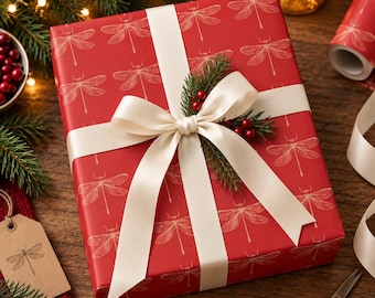 May include: A rectangular gift box wrapped in red paper featuring a repeating white dragonfly pattern. The box is tied with a cream-colored satin ribbon, accented with a small sprig of evergreen needles and two clusters of red berries.