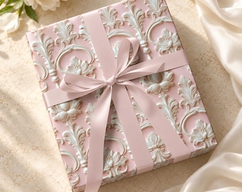 May include: A square gift box wrapped in light pink paper featuring a raised white embossed floral and scroll pattern. The box is tied with a smooth, matching light pink satin ribbon tied in a centered bow across the top surface.