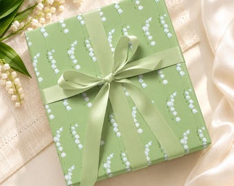 Può includere: Una scatola regalo quadrata avvolta in carta verde chiaro con un motivo ripetuto di fiori bianchi di mughetto. Il pacchetto è legato con un nastro di raso verde chiaro liscio e coordinato, annodato a fiocco al centro della confezione.