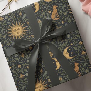 Puede incluir: Una caja de regalo envuelta con un fondo azul oscuro adornado con soles, lunas, zorros y motivos florales. Est&aacute; atada con una cinta y un lazo gris oscuro, creando una presentaci&oacute;n decorativa y elegante.