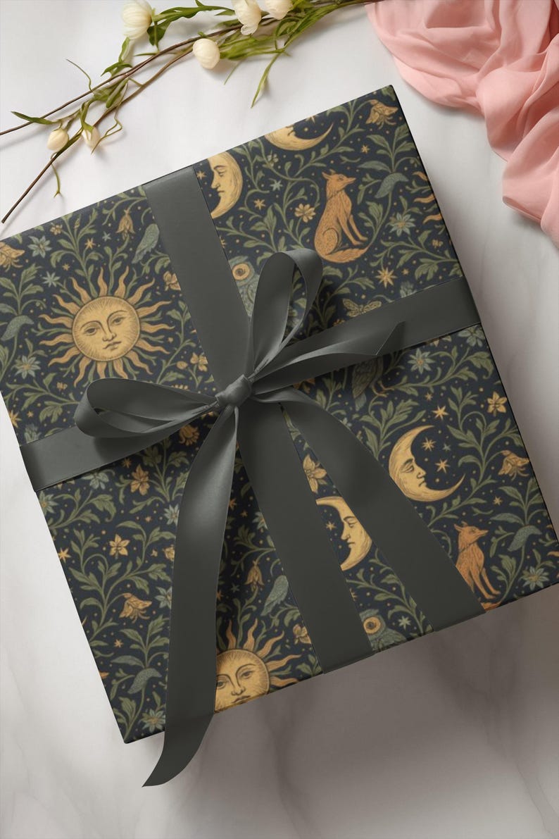 Puede incluir: Una caja de regalo envuelta con un fondo azul oscuro adornado con soles, lunas, zorros y motivos florales. Est&aacute; atada con una cinta y un lazo gris oscuro, creando una presentaci&oacute;n decorativa y elegante.