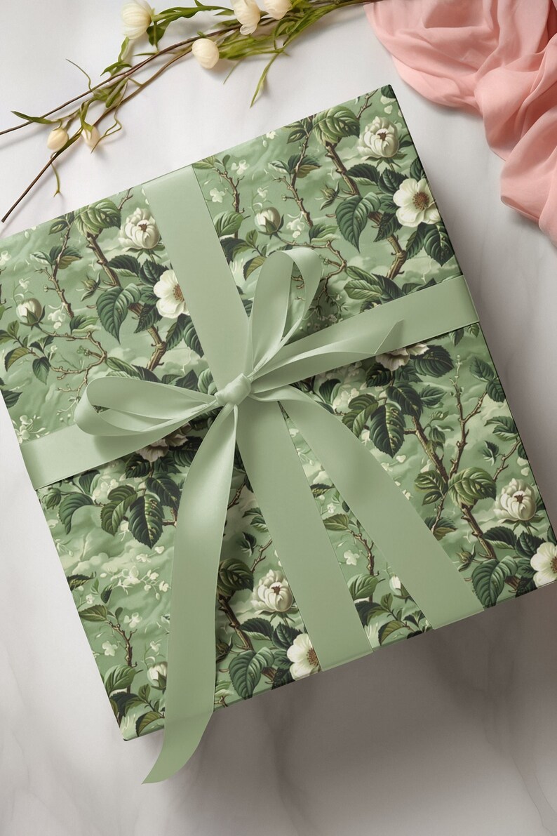 May include: A square gift box wrapped in sage green paper featuring a detailed botanical pattern of white flowers, green leaves, and brown branches. The box is tied with a wide, smooth, sage green satin ribbon arranged in a centered bow.