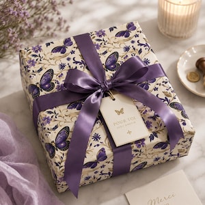 May include: A wrapped gift box with a purple satin ribbon and a tag that reads "Pour Toi Avec Amour." The gift wrap features a cream background with purple butterflies and floral patterns. A lit candle and a small plate are in the background.