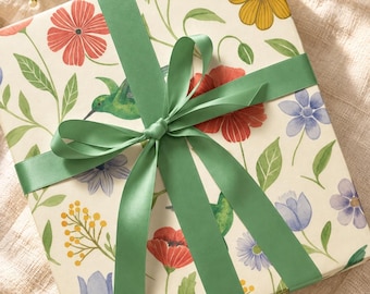 Può includere: Una scatola regalo quadrata avvolta in carta color crema con una stampa botanica colorata di fiori rossi, gialli e blu, foglie verdi e un piccolo colibrì verde, legata con un ampio nastro di raso verde liscio e tinta unita.